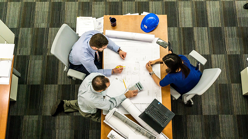 Bilden visar ett arbetsbord uppifrån med fyra personer som arbetar med en byggritning 